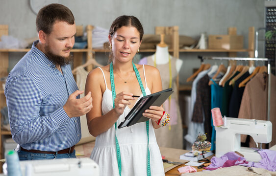 Careful young female dressmaker discussing dress design on tablet with man in fashion studio