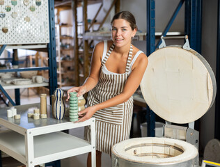 Young potter girl takes out finished ceramic dishes, various mugs and a vase from the oven