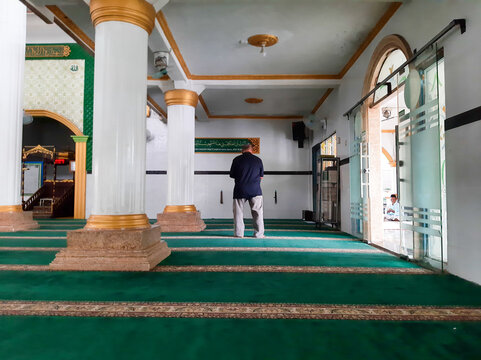 Man praying inside a mosque with green carpet and white pillars.
