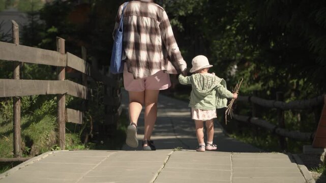 A little girl pink dress carries bundle sticks, she takes her mother's hand look concentration. Shot from behind, family walks along a paved path leading down into the forest on a warm, sunny day.