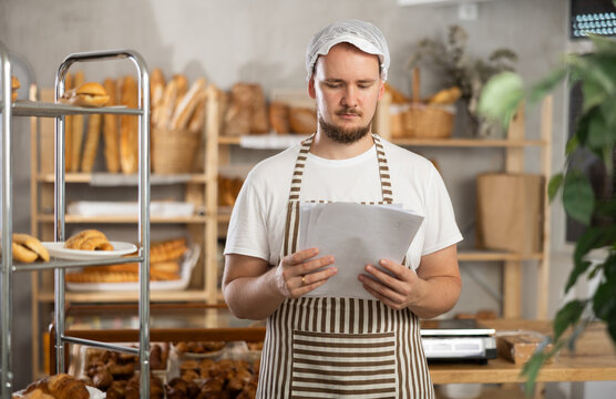 Guy running bakery is standing with papers in hands in middle of bakery. Man manager in apron checks bill of lading, analyzes data in documents