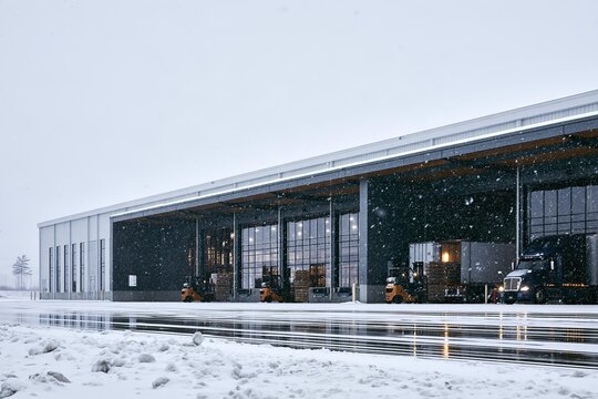 Forklifts move cargo at loading docks. Trucks wait for delivery at industrial warehouse. Winter snow falls on distribution center