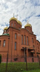 Red brick Orthodox church with golden onion domes, arched windows, ornate crosses, wrought iron fence, and dramatic cloudy sky, exterior architectural photograph © almal