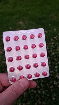 Hand holding pink blister pack with twenty five round tablets against textured green grass, visible pharmaceutical packaging and medicine dosage form outdoors