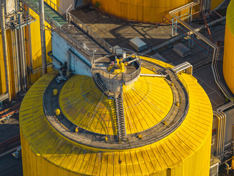 Aerial view of an industrial anaerobic digester with yellow tanks, metal pipelines, access platforms, and processing equipment at a wastewater treatment plant in Italy.