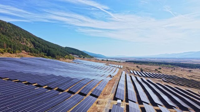 Solar panels are positioned in long rows across a large field near a mountain range under clear skies. The landscape shows a blend of technology and nature.