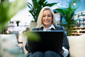 Obraz premium Professional businesswoman types on a laptop from a cushioned rooftop lounge surrounded by greenery, illustrating concentrated remote work in a comfortable urban outdoor workspace.