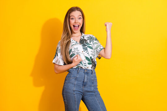 Stylish young woman celebrating success with cheer over vibrant yellow background wearing trendy print shirt and denim