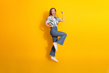Fototapeta premium Cheerful young woman in fashionable casual attire jumping with joy on yellow background showcasing vibrant lifestyle