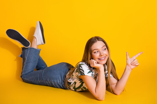Lovely cheerful young woman pointing in a playful manner while lying on a vibrant yellow background in stylish attire