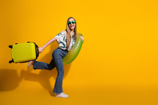 Charming young woman posing with a green suitcase and pool float on a cheerful yellow background, ready for vacation