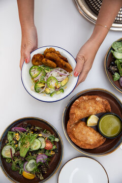 overhead shot of female hands putting a dish of fritters and veggies on to a white table with samosas and a salad