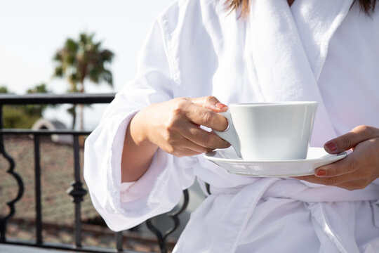 woman in white plush hotel robe with a cup of coffee in hand outdoors on a hotel patio morning