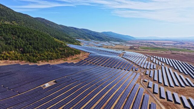 Large solar panel farm spreads across rolling hills in a rural area. Solar panels align in rows, soaking up sunlight. Mountains stand in the background under a clear blue sky.