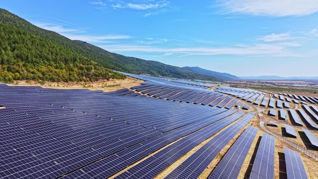Solar panels stretch across a vast field next to mountains. The sky is clear and blue, showing bright daylight. The scene illustrates renewable energy efforts.