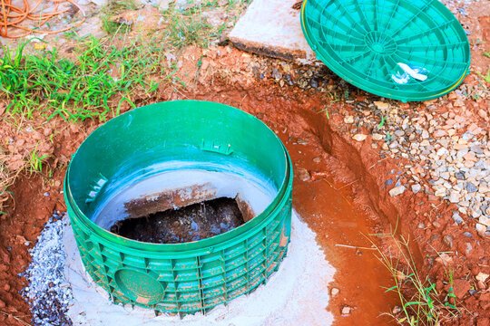 Septic tank plastic sewer access cover is open in dug area with soil, gravel nearby.