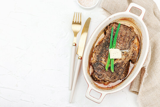 Raw ribeye steak in ceramic baking dish with herbs, spices, cutlery and napkin on white plaster