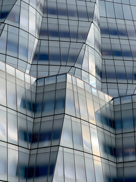 Abstract view of a modern glass building with faceted surfaces reflecting sky and surroundings, forming a geometric pattern of light, shadow, and distortion in an urban environment.