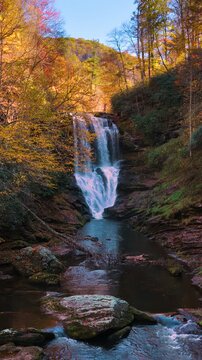 Fall season transforms forest waterfall into vibrant natural attraction. Water flows over rocky cliffs while visitors walk beneath surrounded by golden foliage.