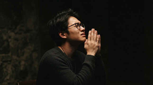Low key dramatic side portrait photo young asian man glasses clasped hands fervent deep prayer gesture dark moody studio background sorrow repentance hope spiritual faith photo