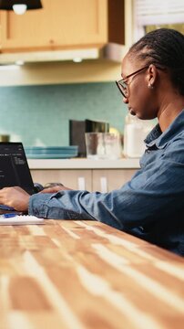 Vertical composition: Person codes on laptop in kitchen during remote IT work, writing source code and using command-line tools for software development and engineering at home.