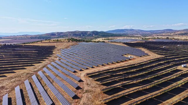 A large field is filled with solar panels arranged in rows under a clear sky. The setup aims to generate renewable energy in a rural area surrounded by hills.