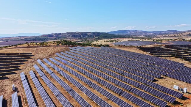 Rows of solar panels cover a dry landscape under clear blue skies. Mountains are visible in the distance. The setting is in a rural area with no signs of urban development.