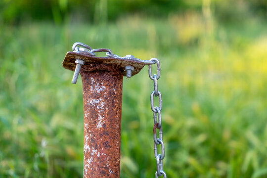 A corroded metal pole topped with a bolted plate holds a metal chain outdoors, surrounded by blurred green grass. The rust and weathered hardware suggest neglect and age.