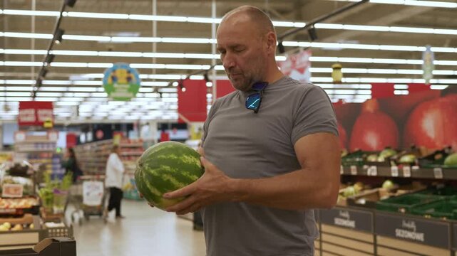 Man in gray shirt examines watermelons in a supermarket, focusing on quality while surrounded by colorful fruits and vegetables in the produce section