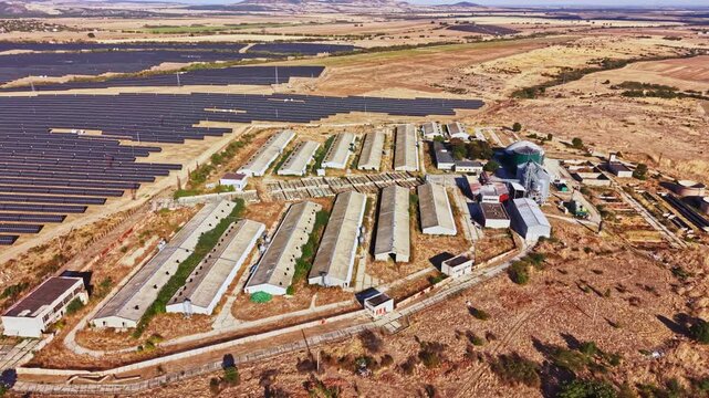 Aerial view of a solar farm next to agricultural buildings in a dry area. There are rows of solar panels alongside a series of structures. The land is mostly clear with some patches of vegetation.
