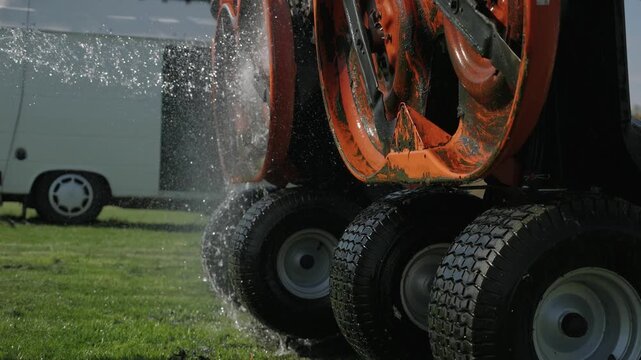 A close-up shot of a jet of water from a hose washing the underside blades of big industrial lawnmowers with water dripping down over the machine onto the tires.