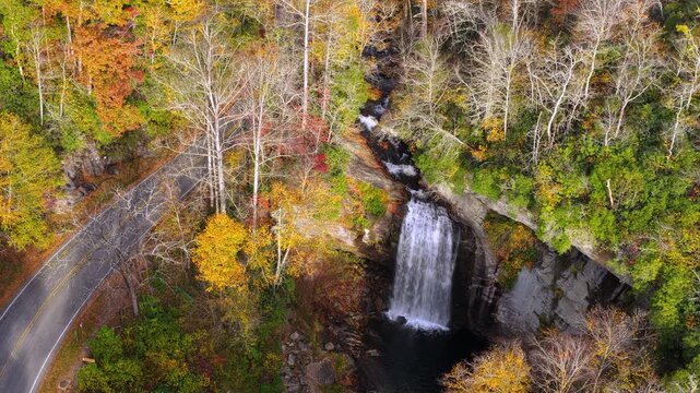 Scenic aerial view of forest waterfall beside quiet roadway. Moving cars and colorful autumn trees create beautiful travel landscape.