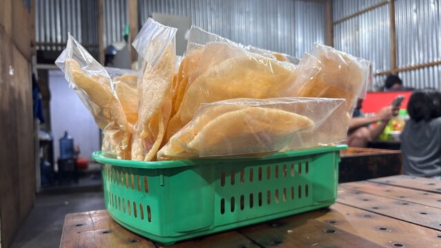 Packaged Indonesian kerupuk prawn cracker snacks in clear plastic bags inside a green basket on a rustic wooden table at a street food warung with corrugated metal walls and blurred background