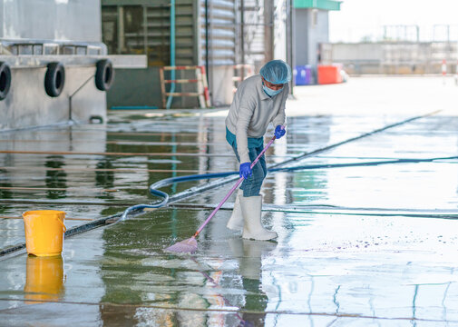 Worker wearing protective gear cleaning outdoor concrete floor with broom and water hose near industrial building.