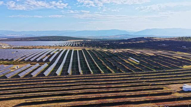 A vast solar farm is visible with hundreds of solar panels aligned in neat rows. The setting features a mountain range in the distance and a clear sky. This scene takes place during daytime.