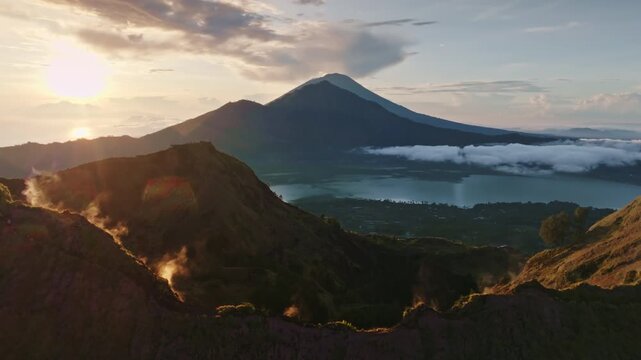 Mount Batur volcano caldera at sunrise, featuring active steam vents on the slopes, shimmering lake, and vibrant sky over surrounding morning landscape. Travel and explore Bali, Indonesia. Static shot
