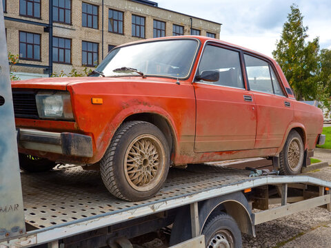 Vintage red Soviet Lada car on a flatbed tow truck platform being transported on a city street with a brick building in background.