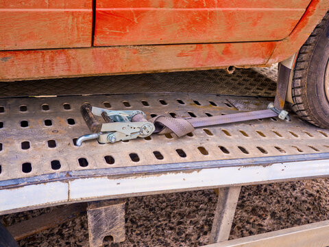 Close-up of heavy-duty ratchet tie-down strap securing a red car wheel on a perforated metal platform of a tow truck or car hauler.