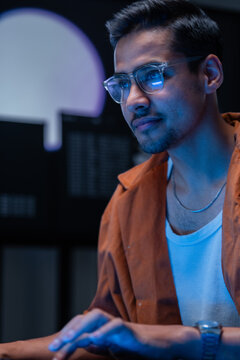 A focused dark-haired cybersecurity analyst works front-facing at the monitor in a blue-lit cyber operations centre, reviewing code and system logs for signs of a cyber attack.