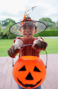 Asian boy in witch hat holding out pumpkin bucket for trick or treat