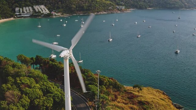 Windmill Viewpoint, wind turbine spinning its blades, generating renewable energy on a lush green hilltop, overlooking turquoise waters of Nai Harn beach in Phuket with many boats moored in bay below