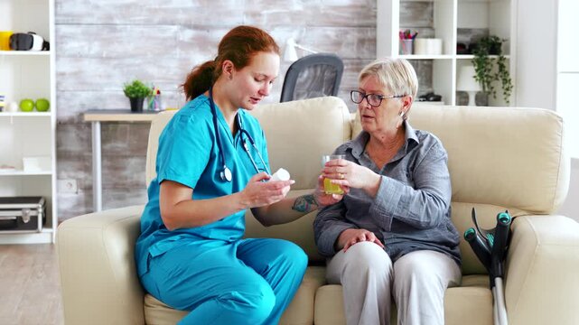Female healthcare worker prepares medication and a drink beside a senior woman, supporting daily medication management, hydration and attentive care.