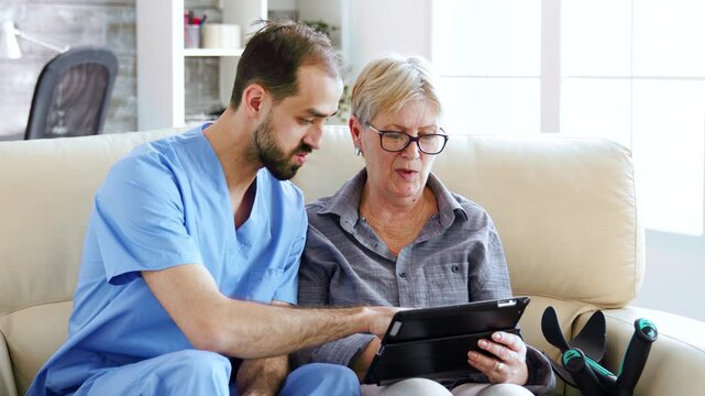 male nurse sits beside a senior woman and guides her on a tablet, representing patient support, digital confidence and assisted learning, further highlighting caregiving and accessibility.