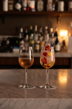 cocktail with floral garnishes and glass of rose wine on a marble bar table in low light with reflections and bar shelf blurred out in background