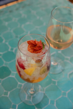 high angle shot of cocktail garnished with florals in a wine glass with straw next to glass of wine on a turquoise tile surface in an ourdoor upscale bar or restaurant patio
