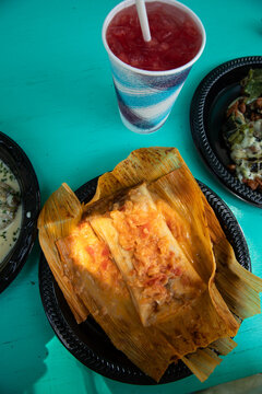overhead shot of traditional mexican tamale wrapped with corn husk on a disposable plate on a bright turquoise table with paper cup of watermelon juice