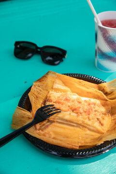 overhead shot of traditional mexican tamale wrapped with corn husk on a disposable plate on a bright turquoise table with paper cup of watermelon juice