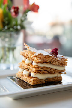 close up image of layers of mille-feuille pastry on white dish in minimal modern cafe with flowers and window in the background