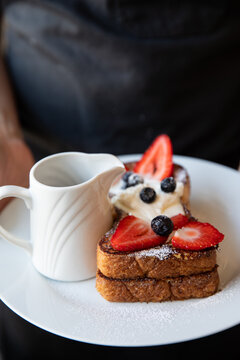 waitress with dark apron holding plate of french toast with fresh berries and cream with a syrup pitcher on a white plate