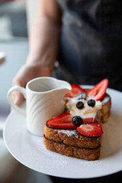 waitress with dark apron holding plate of french toast with fresh berries and cream with a syrup pitcher on a white plate